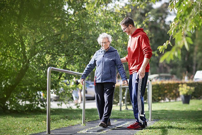 Dies ist eine Patientin, draußen an einem Fitnessgerät, betreut von einem Bewegungstherapeuten.