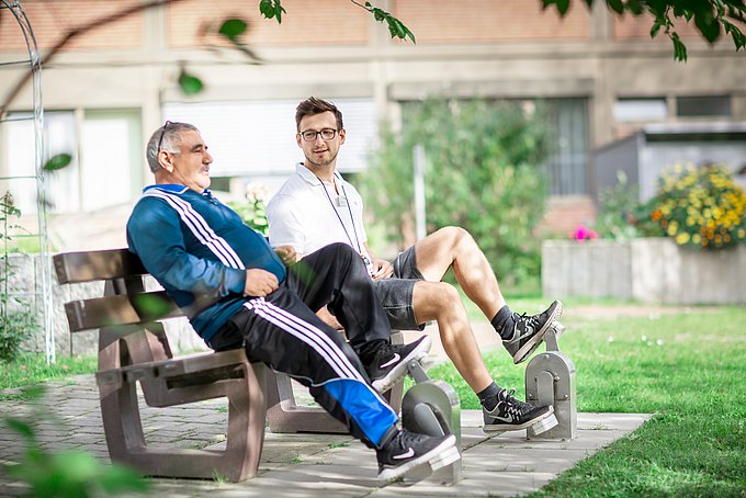 Dies sind ein Patient und ein Trainer an einem Reha-Gerät im Außenbereich der Klinik.